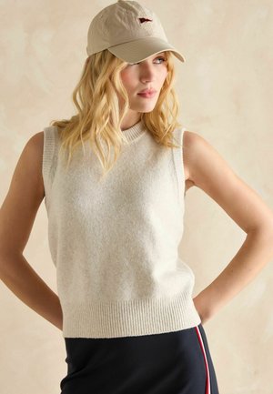 Cremefarbener ärmelloser Pullover mit geripptem Saum und weicher Textur, kombiniert mit einer beige Kappe mit einem kleinen Emblem. Dunkler Rock mit seitlichen Streifen.