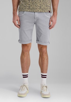 Man wearing light gray rolled-up shorts, white socks with two dark stripes, and beige and white sneakers standing against gray background.