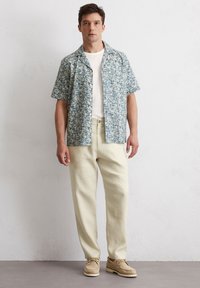 Korte mouwen overhemd met blauw bloemenpatroon, crèmekleurig t-shirt eronder, lichtbeige broeken en tan schoenen met veters. Zachte stoftextuur.
