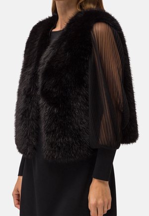 Chaleco de piel sintética negra llevado sobre un vestido negro con mangas largas transparentes y plisadas, mostrado en una persona desde los hombros hasta las caderas.