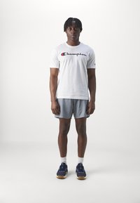 Camiseta blanca de algodón con el logo de "Champion", pantalones cortos de malla gris y zapatillas deportivas azul marino con suelas de color claro, de pie frente a un fondo liso.