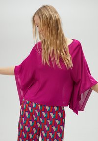 Haut oversized fuchsia à manches courtes et larges, fabriqué en tissu léger. Assorti à un pantalon à motifs présentant des formes géométriques aux couleurs vives.