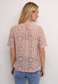 Blusa in pizzo rosa con un motivo geometrico, maniche corte e chiusura con apertura a chiave sul retro. Texture con dettagli a intaglio e bordi smerlati.