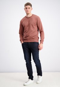 Sudadera marrón con cuello redondo y puños acanalados, combinada con jeans azul oscuro y zapatillas blancas. Textura suave, corte casual.
