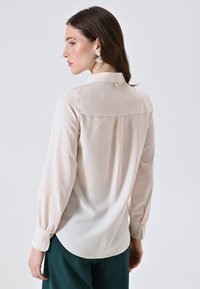 Blusa rosa chiaro con colletto abbottonato, maniche lunghe a sbuffo e orlo arrotondato. Tessuto morbido e liscio con una leggera lucentezza.