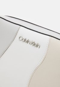 Torebka Calvin Klein w gładkim, białym i beżowym kolorze z czarnymi akcentami, tłoczonym logo i zakrzywionym szwem.