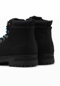 Botas de tobillo de ante negro con cordones azul, cuello acolchado, ojales de latón y suela de goma negra texturizada. Vista trasera que muestra detalles de la construcción del zapato.