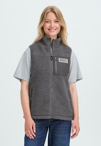 Femme souriante portant un gilet en polaire gris avec des poches zippées sur la poitrine et sur les côtés, sur un t-shirt gris et un jean bleu, devant un fond uni.