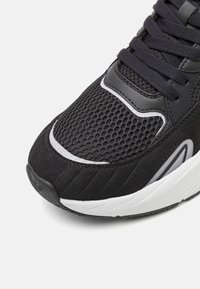 Fekete sportcipő, hálós felsőrésszel, velúr kiegészítőkkel, gumitalppal, fehér részletekkel és textil fűzővel. Közeli nézet az orr részről.