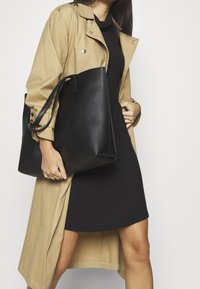 Schwarze Ledertasche mit weicher Textur und langen Trägern, getragen von einer Person in einem beigefarbenen Trenchcoat über einem schwarzen Kleid.