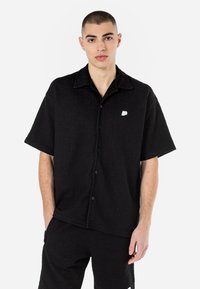 Black short-sleeve button-up shirt featuring a textured fabric, a collar, and a white logo on the left chest. Paired with matching pants.