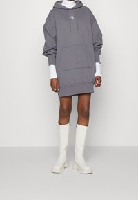 Grå oversized hoodie-klänning med framficka och logotyp, lager på lager över en vit långärmad tröja. Matchad med knähöga vita stövlar.