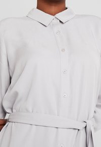 Abito camicia grigio chiaro con collo a camicia, chiusura frontale con bottoni e una cintura coordinata in vita. Tessuto liscio con una leggera lucentezza.
