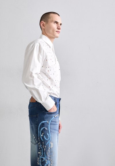Weißes Langarmhemd mit roten und schwarzen Punktverzierungen, kombiniert mit blauen Jeans aus Denim mit dekorativen blauen Nähten und Pailletten.