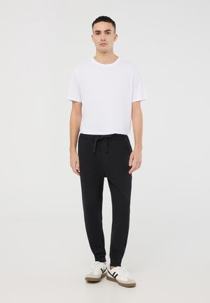 Maglietta di cotone bianca abbinata a pantaloni jogger neri con vita regolabile e polsini elasticizzati. Calzature: sneakers bianche con accenti marroni.