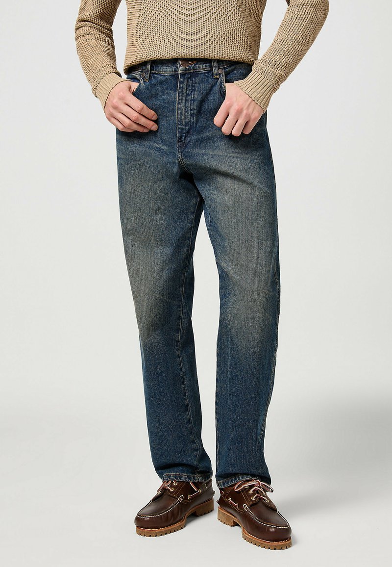 Jeans in denim di un blu scuro con una leggera scoloritura sulla parte anteriore, vestibilità rilassata, design a cinque tasche, indossati con scarpe marroni in pelle con lacci.