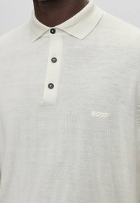 Polo beige clair en tissu texturé, doté d'un col, de trois boutons noirs, et d'un logo blanc subtil sur la poitrine.