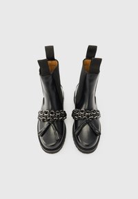 Ζεύγος μαύρων δερμάτινων μποτών μέχρι τον αστράγαλο με παχύ αλυσίδα και λεπτομέρειες με φούντες, ελαστικά πλαϊνά τμήματα και θηλιές έλξης σε άσπρο φόντο.