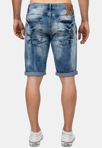Homme portant un short en denim bleu retroussé et des sneakers Converse blanches, se tenant avec les mains détendues le long du corps, vu de dos.