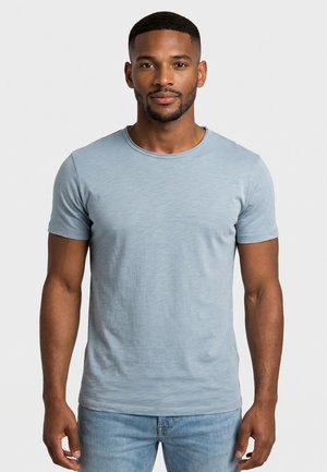 Uomo con capelli corti e barba, indossa una maglietta a girocollo azzurro chiaro e jeans blu, in piedi davanti a uno sfondo chiaro uniforme.