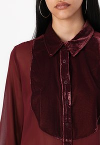 Burgundska bluza z lesketavim teksturiranim prednjim delom, ki ima klasičen ovratnik in zapenjanje na gumb, dopolnjena s polprosojnimi rokavi.