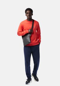 Felpa rossa, pantaloni blu navy e sneakers nere indossati da una persona che tiene una borsa nera con un motivo geometrico e una tracolla.