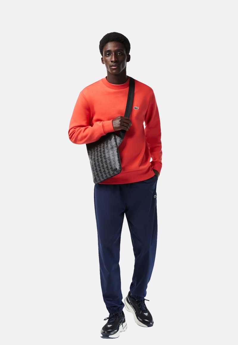 Czerwona bluza, granatowe spodnie i czarne trampki noszone przez osobę trzymającą torbę w geometryczny wzór w kolorze czarnym z paskiem na ramię.