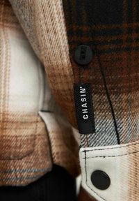 Camisa de cuadros en colores marrón, negro y crema; presenta un botón negro, costuras blancas y una etiqueta que muestra "CHASIN’". Textura suave.