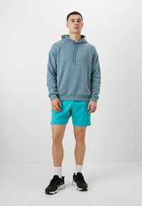 Svetlomodrá mikina s kapucňou a šnúrkami, s logom Nike, v kombinácii s jasne tyrkysovými šortkami a čiernymi športovými topánkami. Viditeľné biele ponožky.