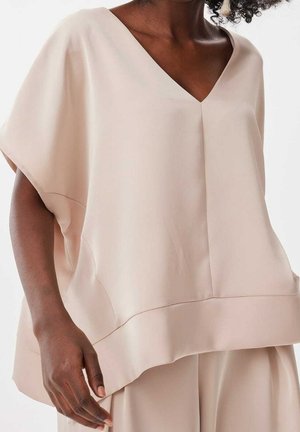 Vrouw draagt losse, lichtbeige V-hals top met brede korte mouwen en bijpassende broek tegen een effen achtergrond.