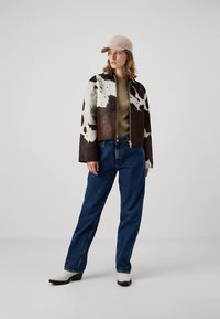 Frau mit beiger Kappe, kurz geschnittenem Jackett mit Kuhmuster, olivgrünem Oberteil, blauer Jeans und weißen Stiefeln, die vor einem schlichten Hintergrund steht.