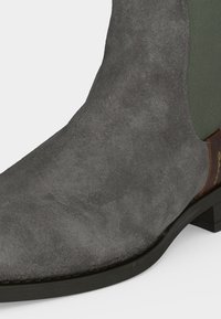 Bottine en daim gris avec semelle noire, panneau latéral élastique et languette de talon marron avec nom de la marque.
