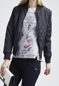 Giacca bomber nera trapuntata con polsini a coste, abbinata a una t-shirt grigia con grafica e jeans scuri. Tiene una borsa a tracolla bianca.