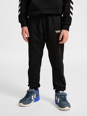 Barn iført sorte Hummel sportbukser og blå sneakers, stående mod en ensartet lys baggrund.