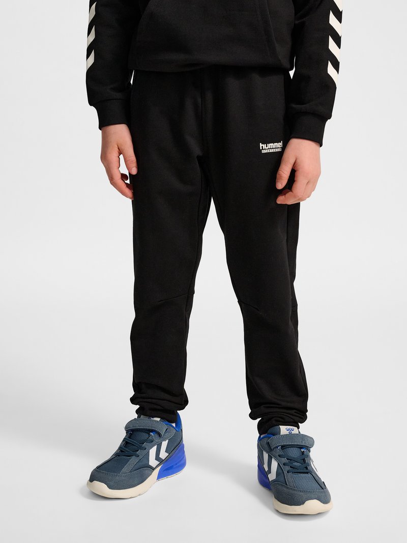 Dziecko w czarnych spodniach sportowych marki Hummel i niebieskich trampkach stoi na jednolitym jasnym tle.