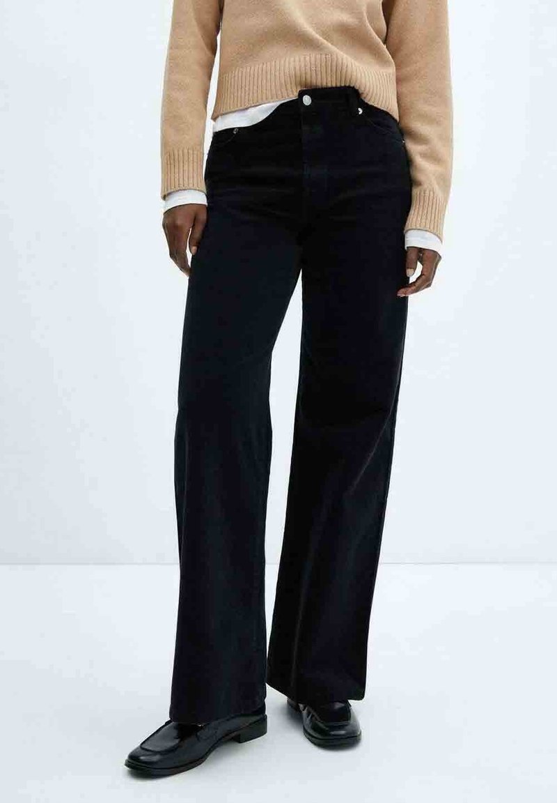 Personne portant un pantalon noir à jambes larges, des mocassins noirs, un pull beige et une chemise blanche sur un fond clair uni.