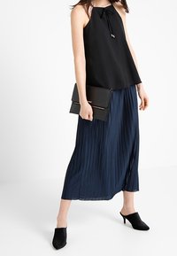 Svart ärmlös blus med en rosett vid halsen, tillsammans med en plisserad marinblå maxikjol och svarta klackmules, hållandes en svart kuvertväska.