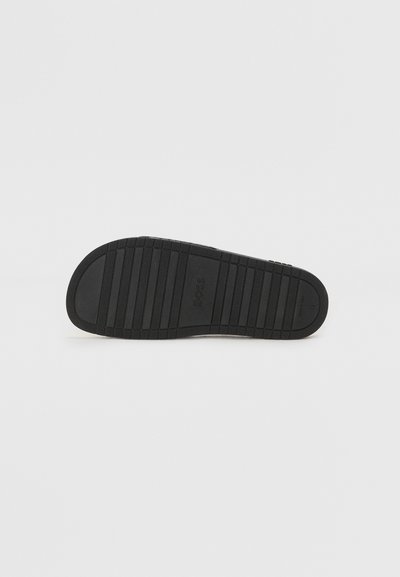 Semelle de chaussure en caoutchouc noir avec un motif de bande de roulement texturé et un logo de marque en relief. Design plat, sans marques visibles sur le dessus.