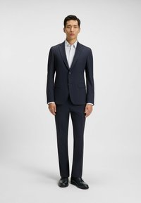 Mörkblå kostym med skräddarsydd passform, kavaj med en knapp, matchande byxor och en vit skjorta med ett ljust prickmönster. Svarta skor.