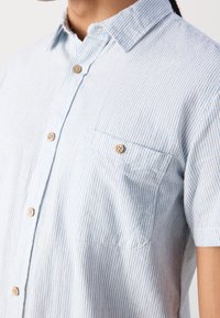 Chemise à manches courtes rayée bleu clair et blanc avec des boutons en bois et une poche poitrine boutonnée.