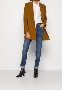 Kamel-farbener, doppelt geknöpfter Blazer über einem weißen Hemd, kombiniert mit blauen Skinny-Jeans. Schwarze, hohe Stiefeletten. Glatte Textur, strukturiertes Design.