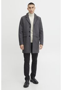 Manteau en laine gris avec revers crantés, fermeture à boutons et poches avant, porté sur un col roulé crème, associé à un pantalon noir et des chaussures noires épaisses.