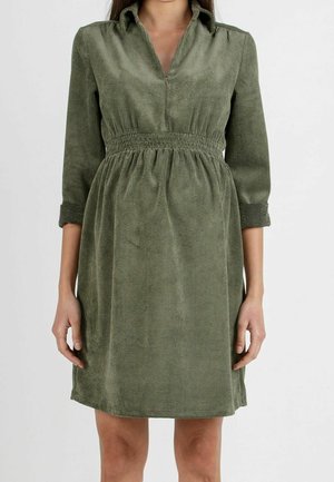 Nainen pukeutuneena polvipituiseen oliivinvihreään corduroymekkoon, jossa on kolme neljäsosakoon hihat ja vyötärölle kerätty vyötärönauha, seisoo yksiväristä taustaa vasten.