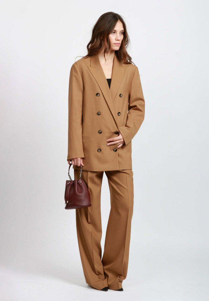 Blazer doppiopetto color cammello con bottoni neri, abbinato a pantaloni a gamba larga dello stesso colore; tiene in mano una piccola borsa bordeaux scuro.
