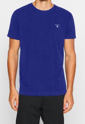 Mann trägt ein einfaches blaues T-Shirt mit kurzen Ärmeln und kleinem Gant-Logo auf der linken Brust sowie schwarze Hose.