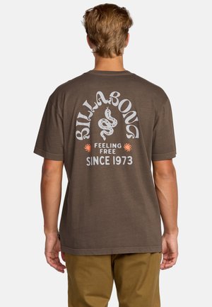 Mężczyzna ubrany w brązowy t-shirt Billabong z białą grafiką węża i napisem „Feeling Free Since 1973” na plecach, w połączeniu z khaki spodniami.
