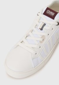 Zapatilla deportiva blanca con una puntera de cuero liso, lados de malla, ojales reforzados y una etiqueta de logo en burdeos en la lengüeta.