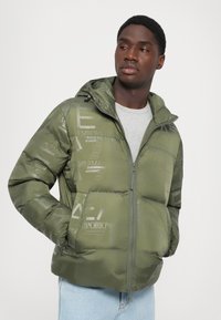 Jachetă de umplutură verde măsliniu cu glugă, cu închidere prin fermoar și imprimeu de branding subtil. Textură netedă și design matlasat, cu buzunare laterale.