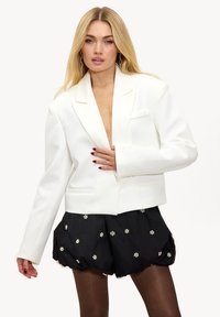 Witte getailleerde blazer met ingesneden revers, gestructureerde schouders en twee voorzakken, gedragen over zwarte shorts met kleine witte bloemen.