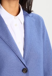 Kvinde iført en blå blazer over en hvid skjorte med knapper, hvor kraven, reverset og én knap er fastgjort på blazeren.